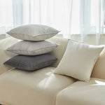 Une pile de coussins gris et blancs, disposés de manière élégante sur un canapé beige, créant une atmosphère apaisante et confortable dans une pièce baignée de lumière naturelle.