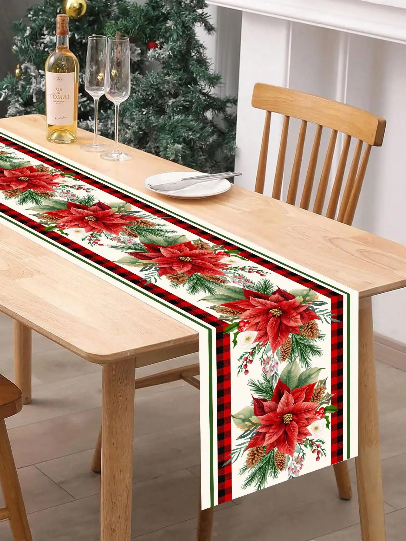 Élégant chemin de table de Noël avec des poinsettias rouges et des branches de sapin sur fond de carreaux noirs et rouges, accompagné d'une bouteille de vin et de verres de champagne sur une table en bois clair.