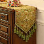 Un magnifique tapis de table orné de motifs floraux dorés et de glands verts décorant avec élégance la commode en bois sombre.