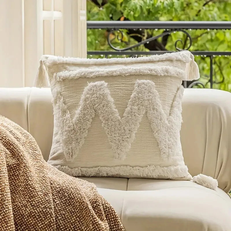 Coussin décoratif moelleux avec des pompons sur un canapé blanc, entouré de plantes verdoyantes visibles par la fenêtre.