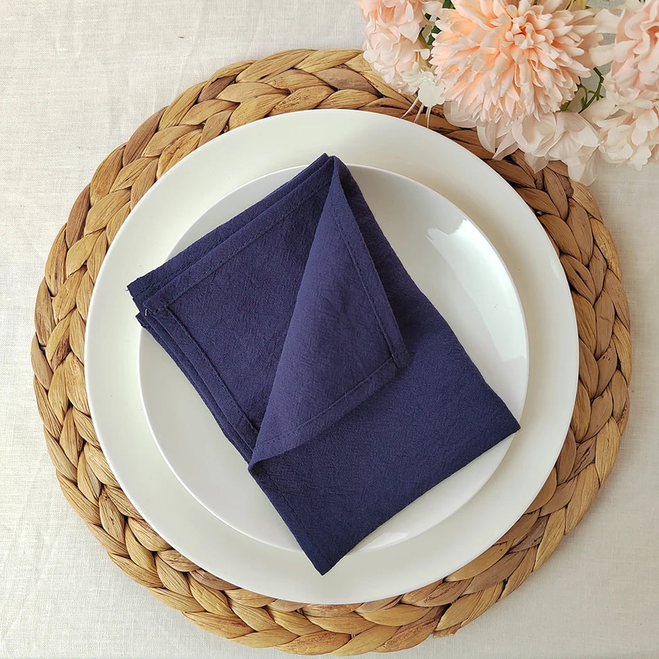 Assiette blanche ronde posée sur un dessous-de-plat tressé en fibres naturelles, avec une serviette de table pliée en forme de triangle sur l'assiette et des fleurs roses sur le côté.