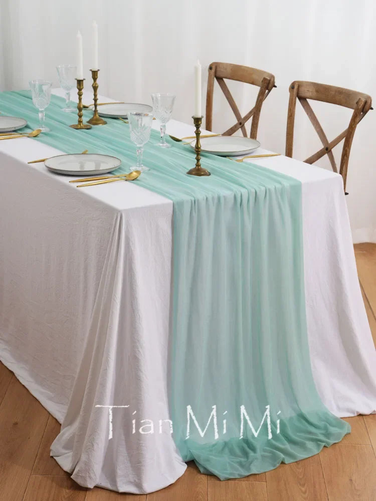La table de salle à manger est magnifiquement dressée avec une nappe blanche et un chemin de table vert menthe, des assiettes et des verres élégants, ainsi que des chandeliers dorés, créant une ambiance chaleureuse et raffinée.