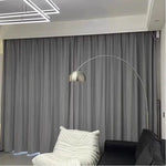 Vue d’ensemble d’un salon moderne avec un rideau gris en velours couvrant entièrement une baie vitrée, aux côtés d’un canapé noir et d’un fauteuil blanc.