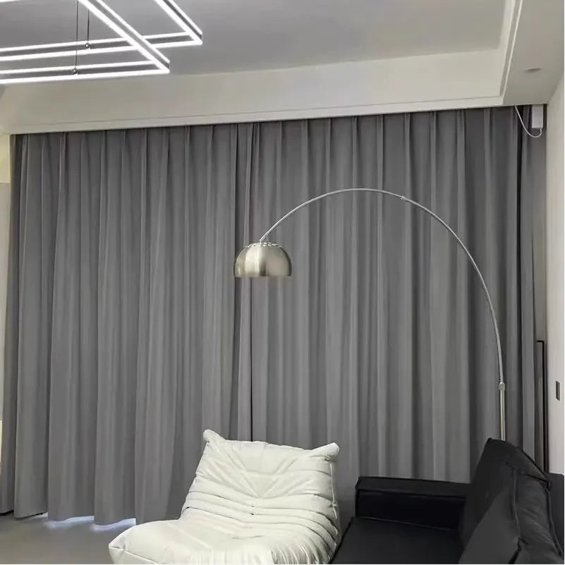 Vue d’ensemble d’un salon moderne avec un rideau gris en velours couvrant entièrement une baie vitrée, aux côtés d’un canapé noir et d’un fauteuil blanc.