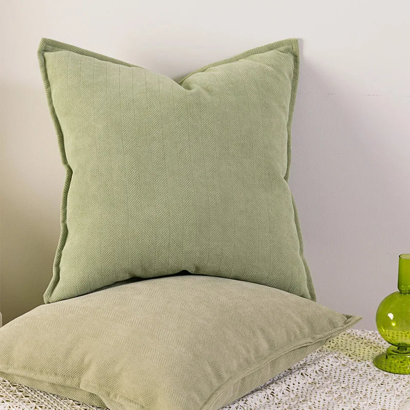 Deux coussins carrés verts pâles aux motifs côtelés, posés sur un tapis blanc moelleux.