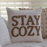 Coussin décoratif gris avec le texte brodé "Stay cozy" en couleur marron, pour une ambiance chaleureuse et confortable dans la chambre à coucher.