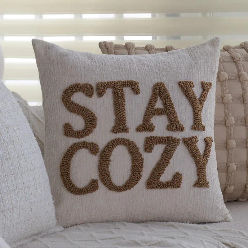 Coussin décoratif gris avec le texte brodé "Stay cozy" en couleur marron, pour une ambiance chaleureuse et confortable dans la chambre à coucher.