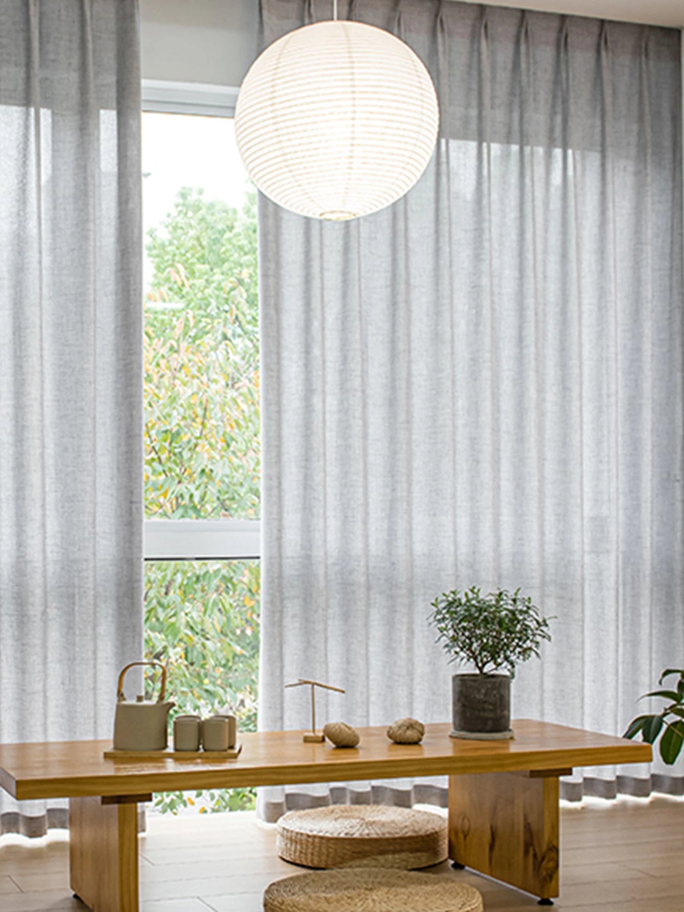 Salon lumineux avec voilage gris clair tamisant, mobilier en bois et suspension en papier.