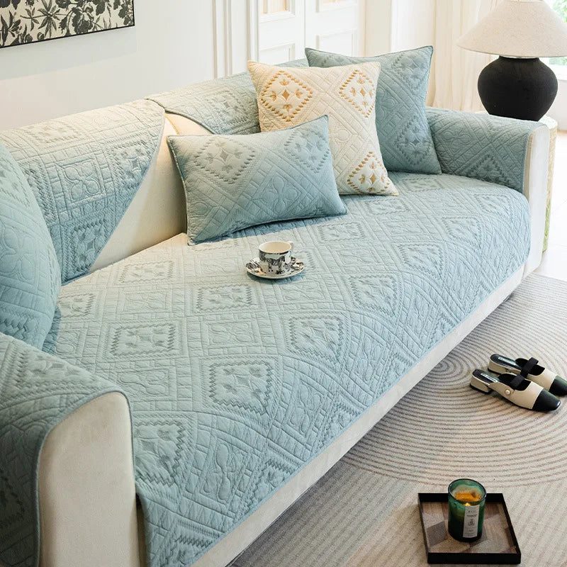 Cet image présente un canapé recouvert d'une courtepointe en couleurs pastel, orné de coussins décoratifs, dans un environnement lumineux et accueillant.