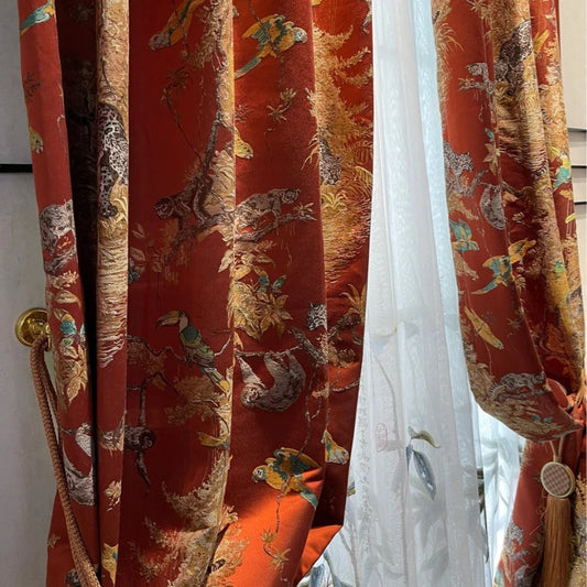 Rideau à motifs tropicaux orangé avec détails animaliers et feuillages dorés, retenu par une embrasse dorée et accompagné d’un voilage blanc délicat.