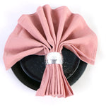 Un napperon en velours rose plié délicatement sur une assiette noire, avec un anneau métallique argenté servant de décoration centrale.