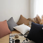 L'image montre un coin de pièce meublé de coussins de différentes couleurs et textures, avec des éléments décoratifs tels que des plumes et un pot, ainsi qu'un ordinateur portable sur un tapis noir et blanc géométrique, créant une ambiance confortable et chaleureuse.