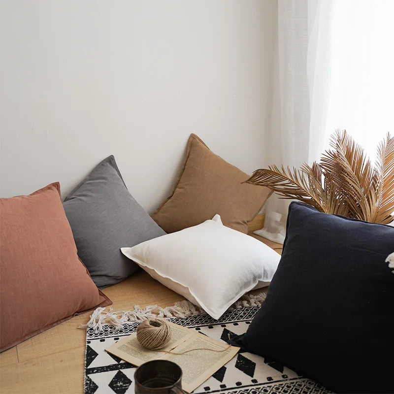 L'image montre un coin de pièce meublé de coussins de différentes couleurs et textures, avec des éléments décoratifs tels que des plumes et un pot, ainsi qu'un ordinateur portable sur un tapis noir et blanc géométrique, créant une ambiance confortable et chaleureuse.