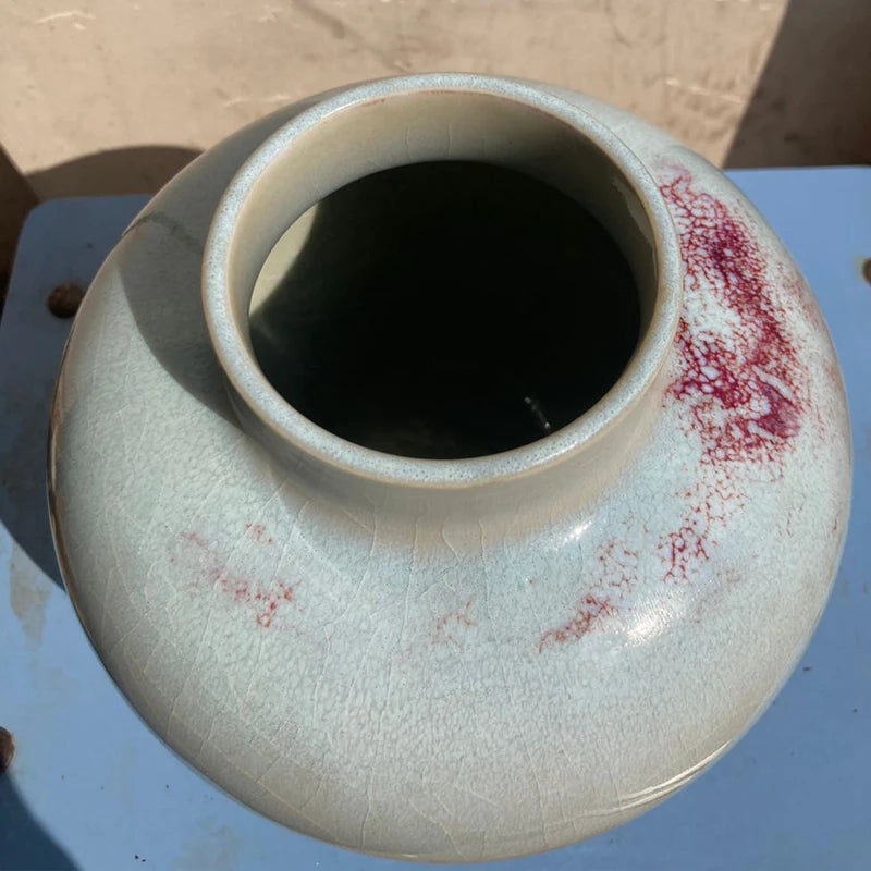 Cette image montre un vase en céramique avec une texture craquelée et des accents de couleur rouge, posé sur une surface bleue.