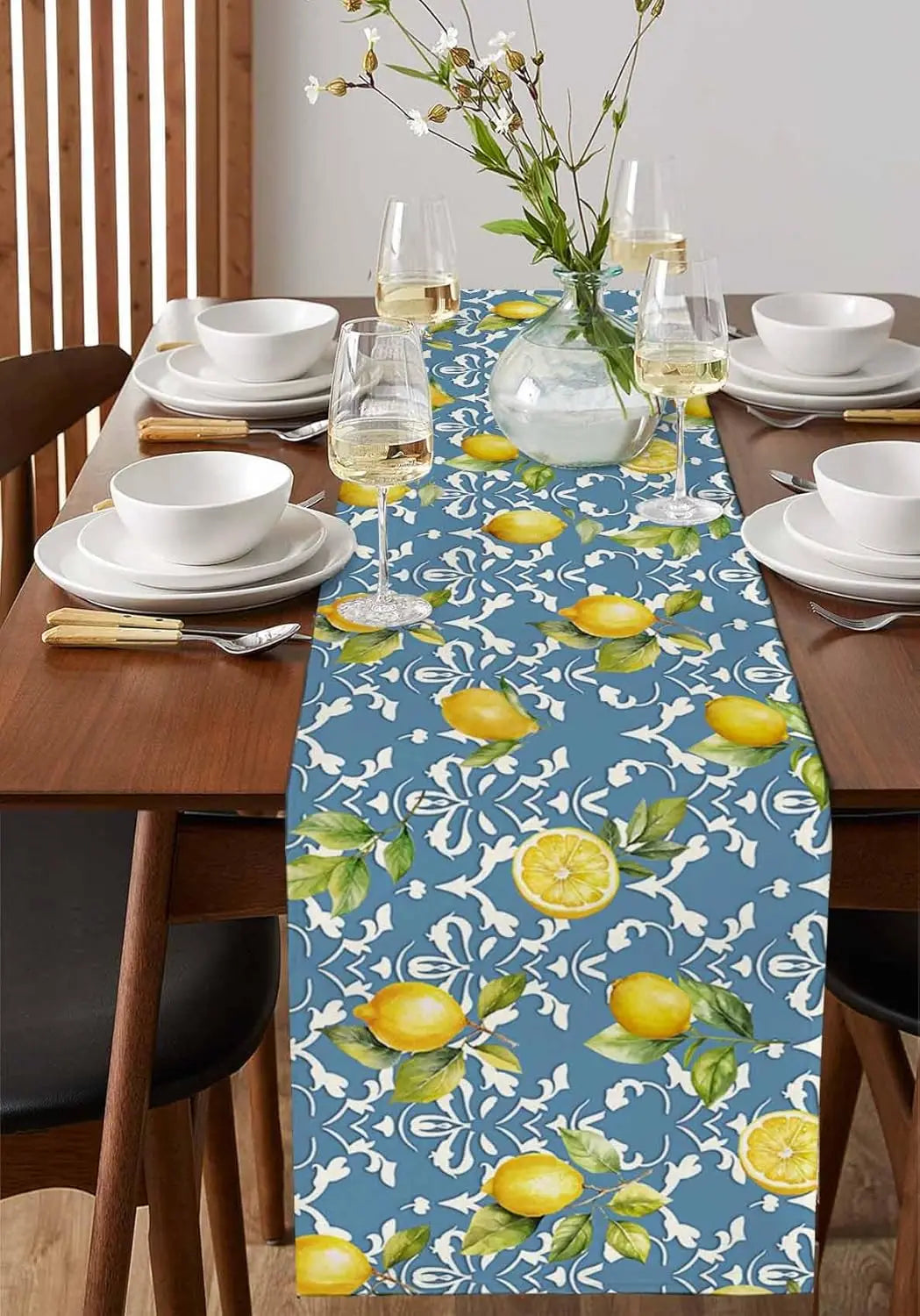 La table est joliment dressée avec des assiettes blanches, des verres à vin et un élégant chemin de table bleu et jaune orné de citrons et de fleurs.