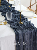 Cette image présente une belle table dressée avec une nappe blanche, une décoration de table élégante composée d'une bougie et d'un dessert dans des assiettes, le tout accompagné d'un magnifique chemin de table gris foncé froissé qui apporte une touche de sophistication à la mise en scène.
