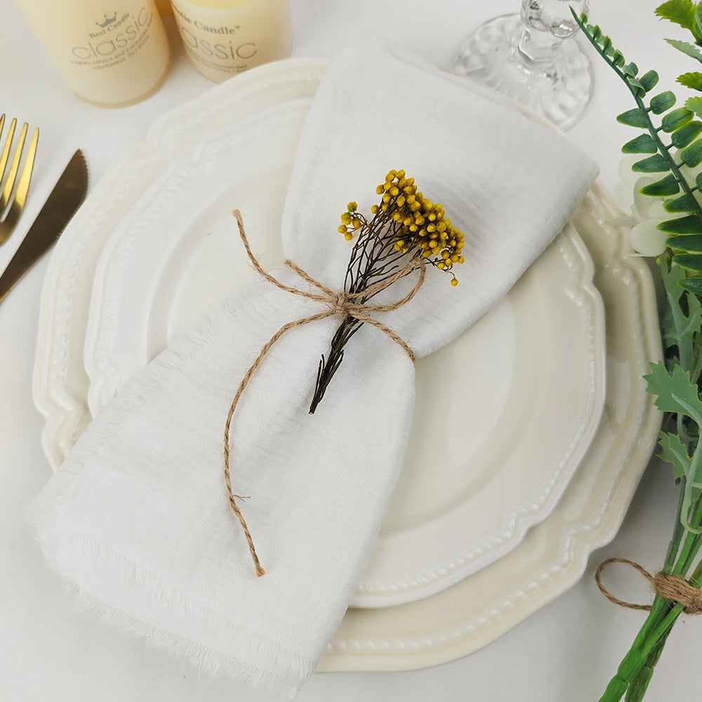 Une serviette blanche avec une petite branche de fleurs jaunes attachée par une ficelle, posée sur une assiette blanche avec d'autres éléments de décoration sur la table.