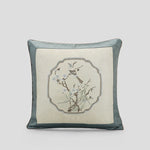 Coussin décoratif avec motif de branche fleurie et oiseau brodé dans un cadre géométrique sur fond ivoire et gris.