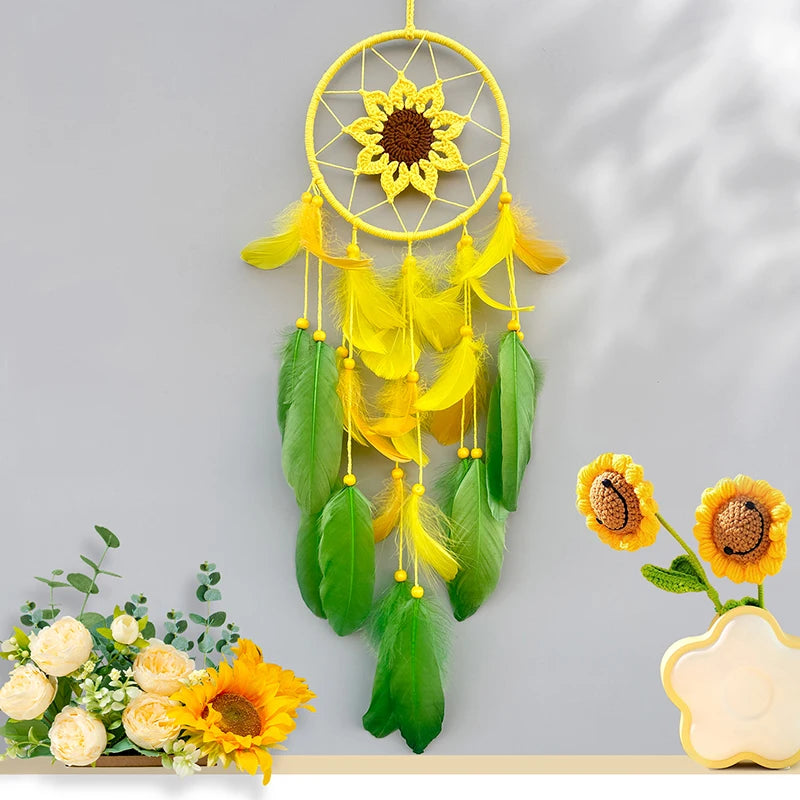 Un capteur de rêves en forme de soleil, décoré avec des fleurs jaunes et des plumes vertes, suspendu sur un mur gris avec un bouquet de fleurs et deux petits tournesols sur une étagère.
