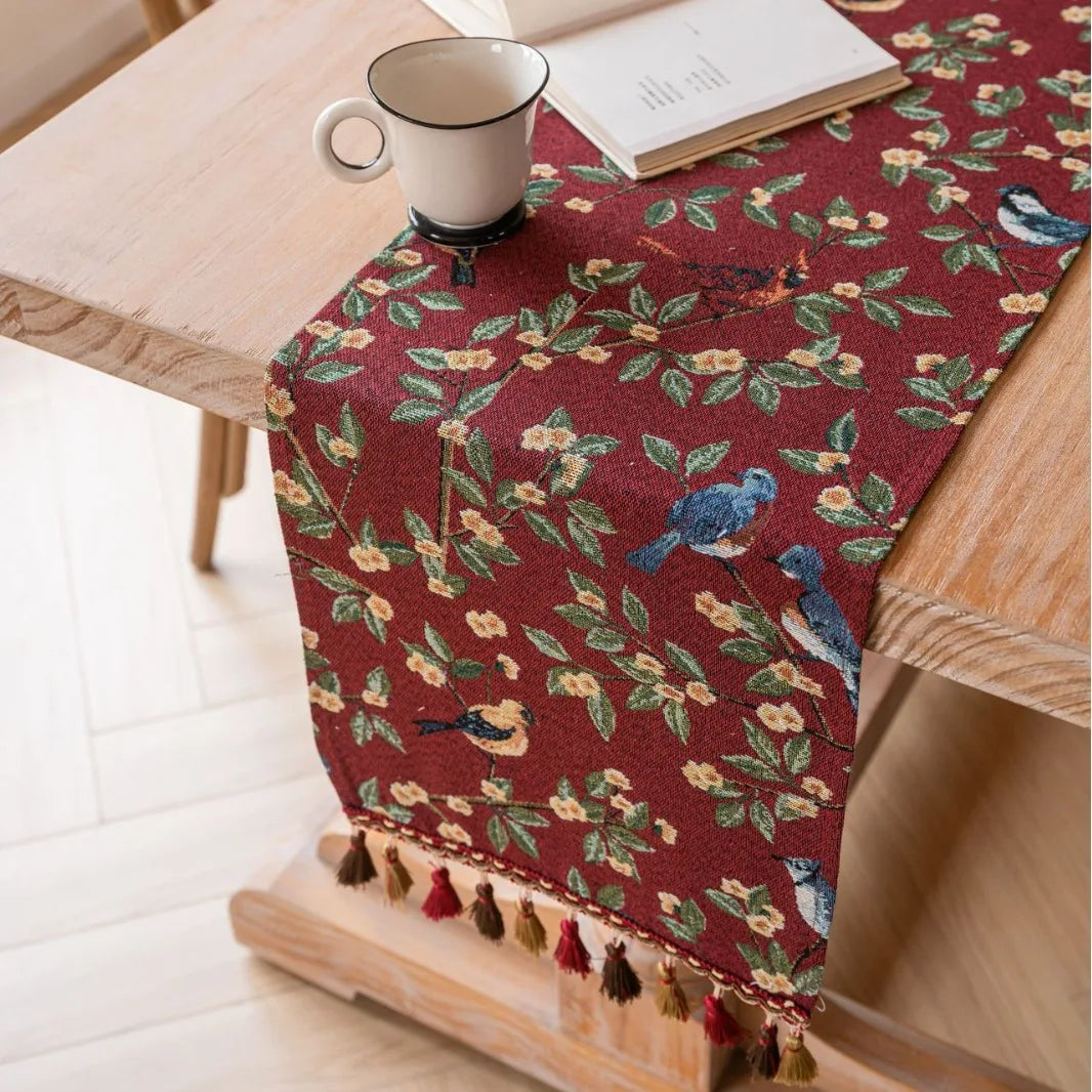 Un élégant chemin de table rouge ornée de motifs floraux et d'oiseaux, sur une table en bois clair, avec une tasse de boisson et un livre posés.