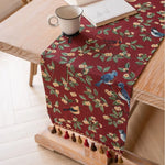 Un élégant chemin de table rouge ornée de motifs floraux et d'oiseaux, sur une table en bois clair, avec une tasse de boisson et un livre posés.