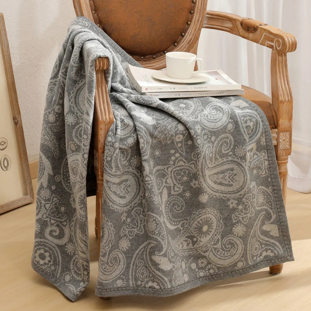 Cette image montre une chaise en bois avec une couverture grise à motifs paisley posée dessus, ainsi qu'une tasse de café et des livres sur l'assise.