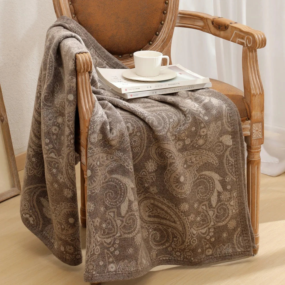 Le fauteuil en bois vintage semble confortable avec sa couverture grise à motifs paisley, posée sur un ensemble de livres et une tasse blanche, créant une ambiance apaisante et cosy.