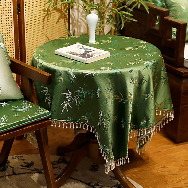 Une table basse ronde couverte d'une nappe verte ornée de motifs de bambous, avec un livre posé dessus et des plantes d'intérieur à l'arrière-plan, créant une atmosphère apaisante et traditionnelle.