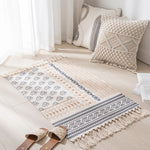Un tapis orné de motifs ethniques avec des franges dans un intérieur lumineux, accompagné de coussins décoratifs et de sandales beiges, créant une ambiance chaleureuse et apaisante.