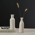 Deux vases blancs sculptés posés sur une surface avec des épis de blé dorés, créant une composition élégante et minimaliste sur un fond noir.