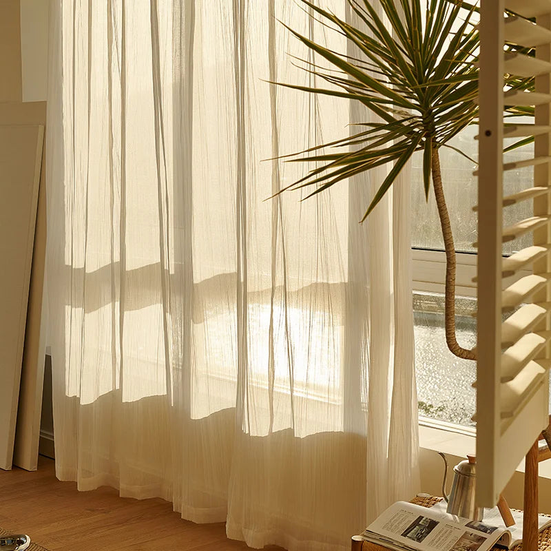 Rideau d'été transparent laissant filtrer les rayons du soleil dans une pièce lumineuse avec plante verte en pot et parquet clair.