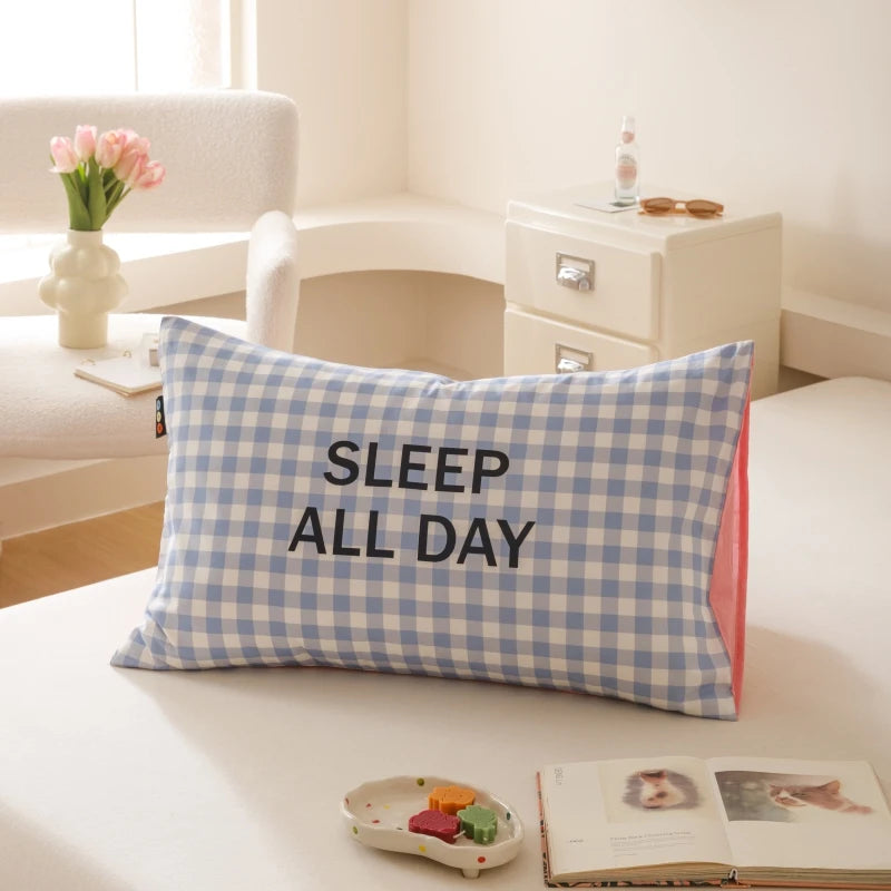 Coussin gris et blanc avec l'inscription "SLEEP ALL DAY" sur une table à côté de fleurs et d'un livre ouvert.