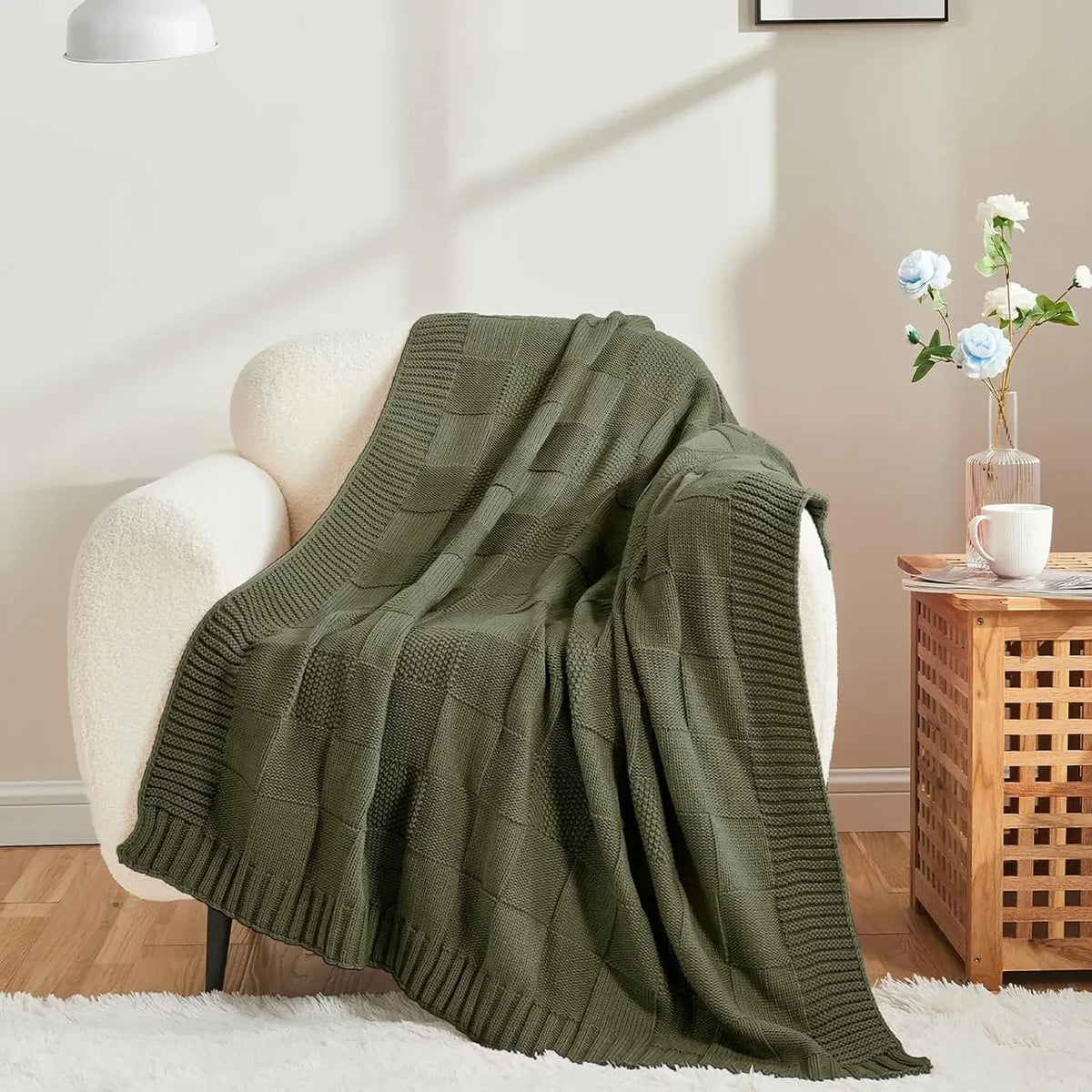 Une couverture tricotée de couleur vert kaki, posée sur un fauteuil confortable avec un vase de fleurs à côté.