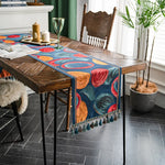 Cette image montre une table en bois rustique avec un chemin de table coloré et décoratif, des bougies, des fleurs et des plantes d'intérieur, créant une ambiance chaleureuse et conviviale dans une pièce lumineuse.