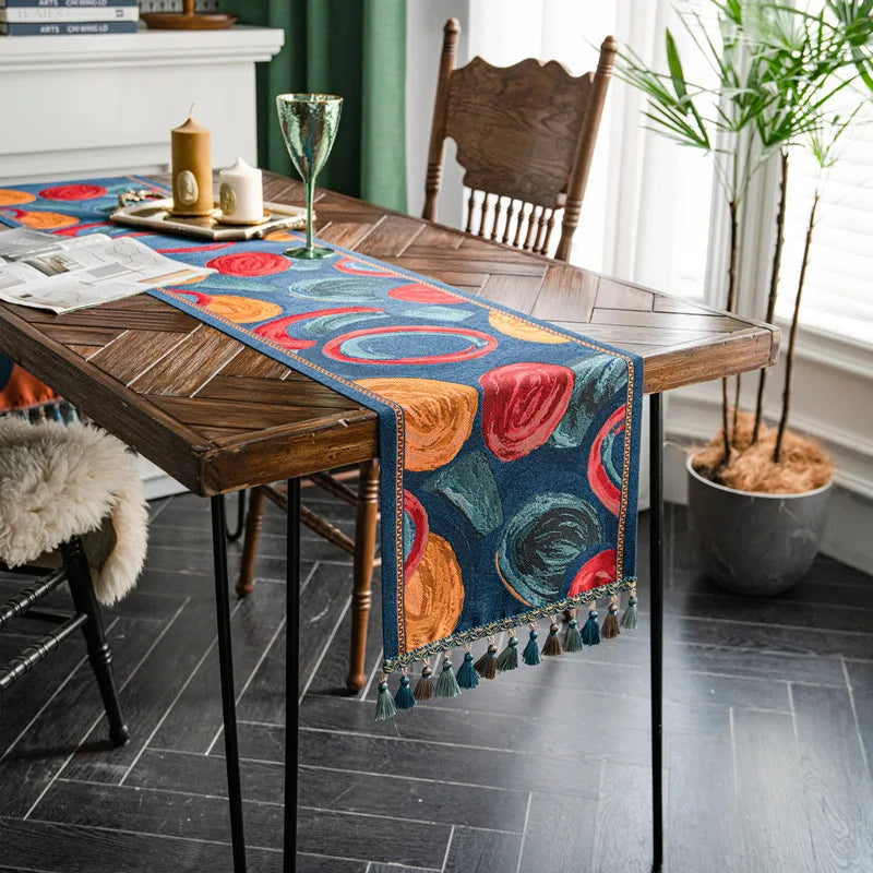 Cette image montre une table en bois rustique avec un chemin de table coloré et décoratif, des bougies, des fleurs et des plantes d'intérieur, créant une ambiance chaleureuse et conviviale dans une pièce lumineuse.