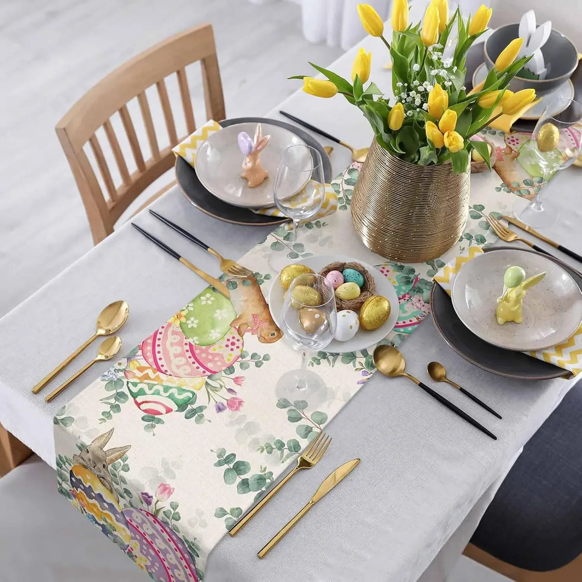 Cette image montre une élégante table de fête printanière, ornée d'un bouquet de tulipes jaunes dans un vase doré, de décorations de Pâques colorées et de couverts dorés sur une nappe fleurie.