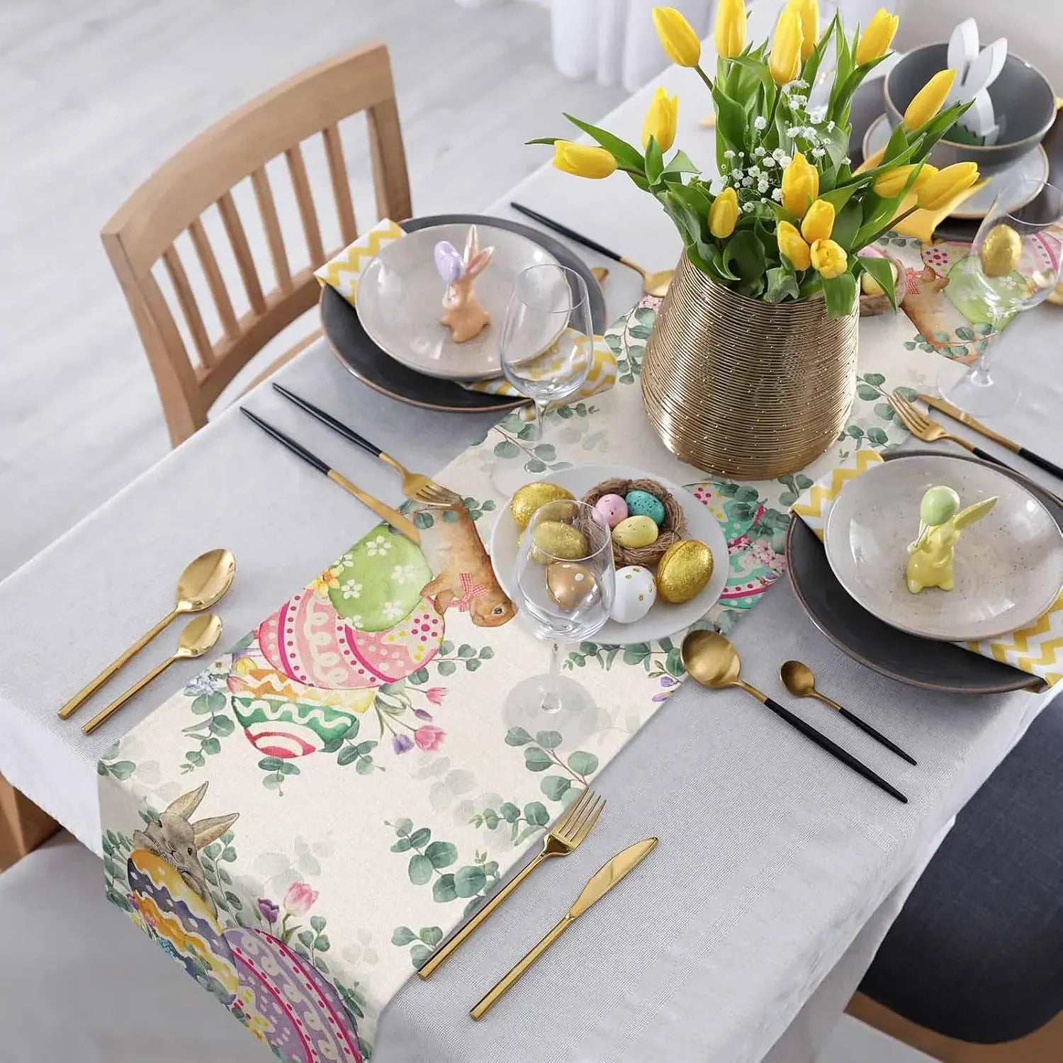 Cette image montre une élégante table de fête printanière, ornée d'un bouquet de tulipes jaunes dans un vase doré, de décorations de Pâques colorées et de couverts dorés sur une nappe fleurie.