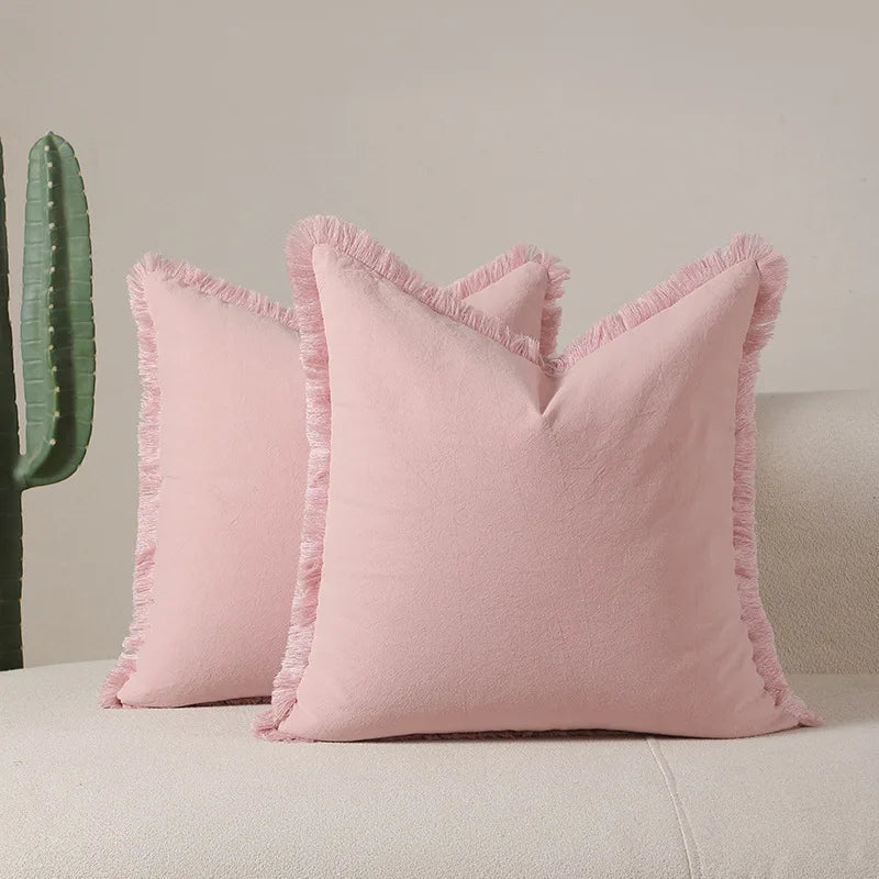 Deux oreillers en velours rose pastel avec des franges sur le pourtour, placés devant un cactus vert sur un sol blanc.