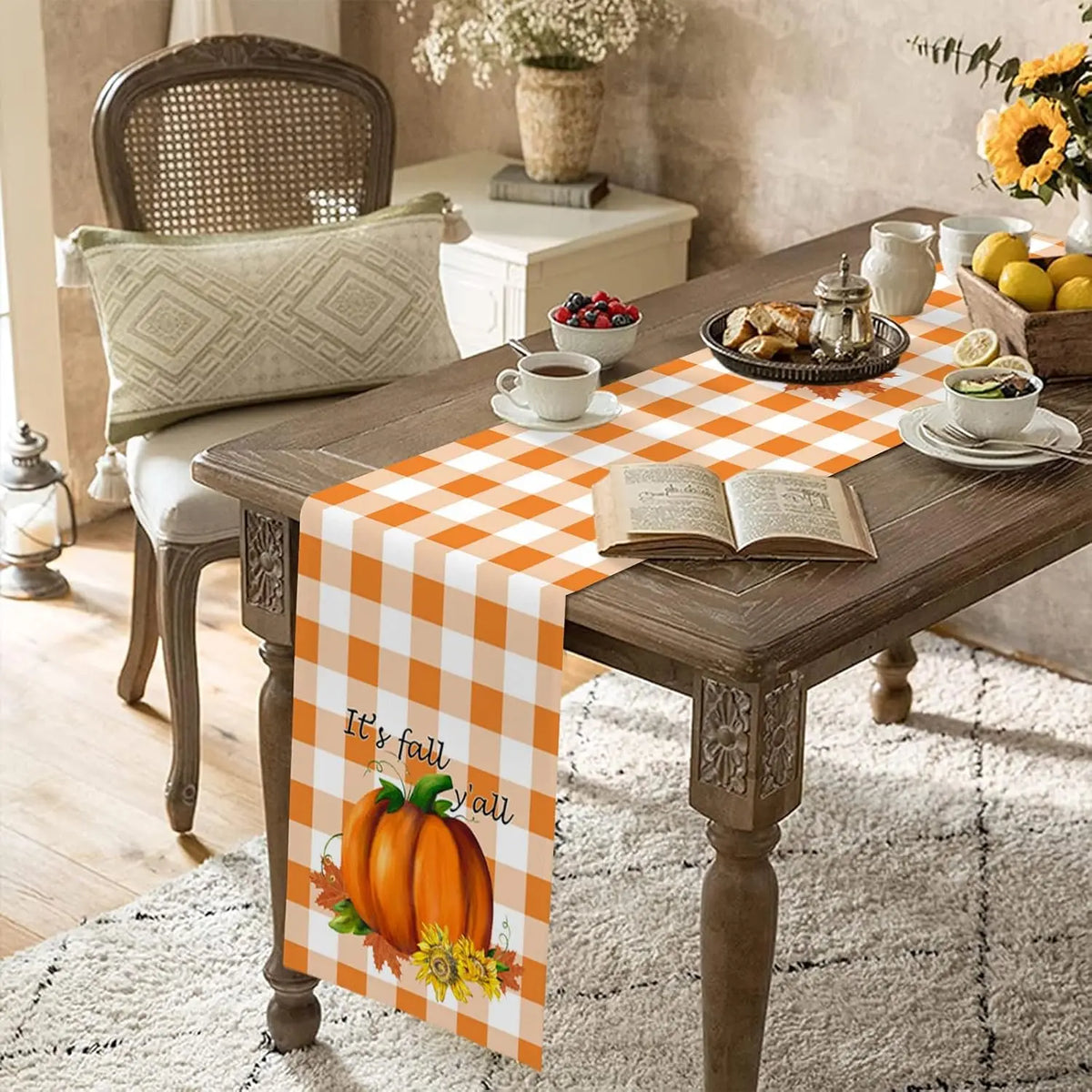 Une table décorée pour l'automne avec un chemin de table à carreaux orange et blancs, une pièce maîtresse de citrouille, des fleurs jaunes et un livre ouvert.