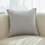 Coussin décoratif gris clair sur un canapé confortable au premier plan, avec un intérieur lumineux et des rideaux blancs en arrière-plan.