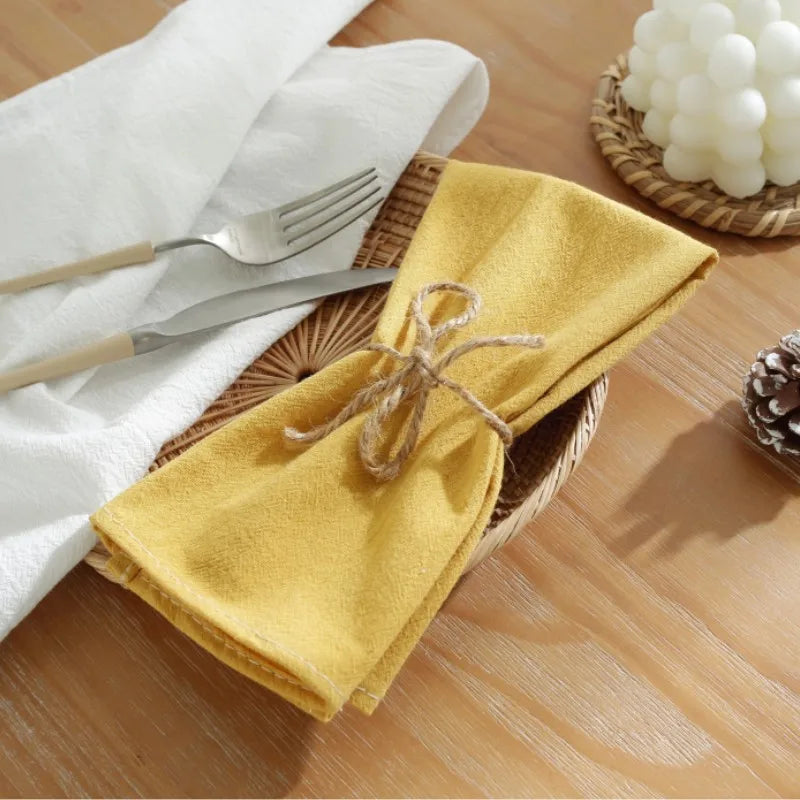 Une serviette de table jaune dorée, délicatement enroulée et attachée avec une ficelle, posée sur une table en bois avec des couverts et une décoration florale, créant une ambiance chaleureuse et accueillante.