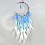 Un capteur de rêves délicat en forme de lune, avec des plumes douces dans des tons de bleu clair et blanc, surmontées de cristaux et de perles scintillantes, suspendu sur un support en bois brun.