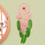 Attrape-rêves rose et vert délicat suspendu sur un mur vert clair avec des fleurs de cerisier en arrière-plan.