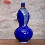 Un vase décoratif en forme de calebasse avec une finition brillante bleu foncé et des touches de rouge, posé sur une surface en bois.