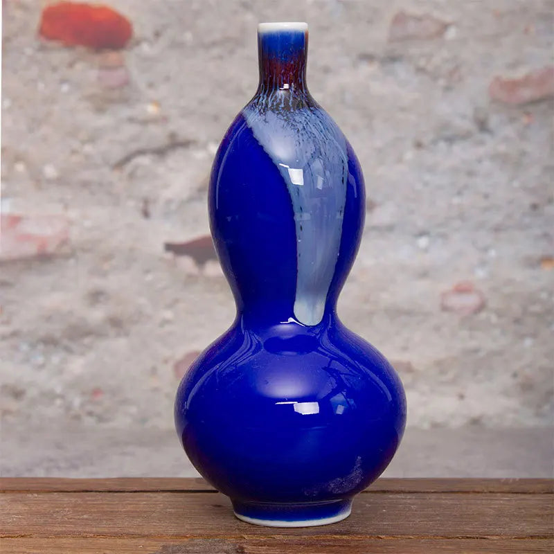 Un vase décoratif en forme de calebasse avec une finition brillante bleu foncé et des touches de rouge, posé sur une surface en bois.
