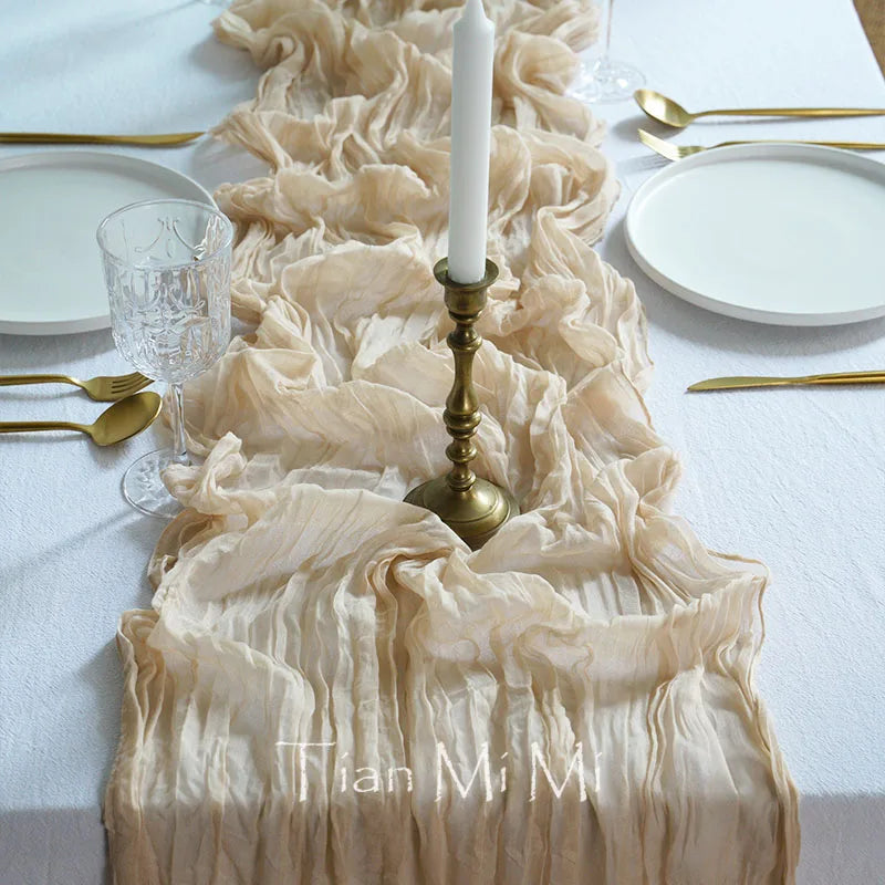 Une magnifique table dressée avec élégance, comprenant une superbe nappe décorative en tissu crème, une bougie blanche dans un chandelier doré, des assiettes et des couverts dorés sur une surface blanche.