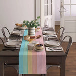 Une scène de table élégante avec une nappe rayée multicolore, de la vaisselle grise, des plantes vertes et un arrangement floral dans un pichet blanc et noir.