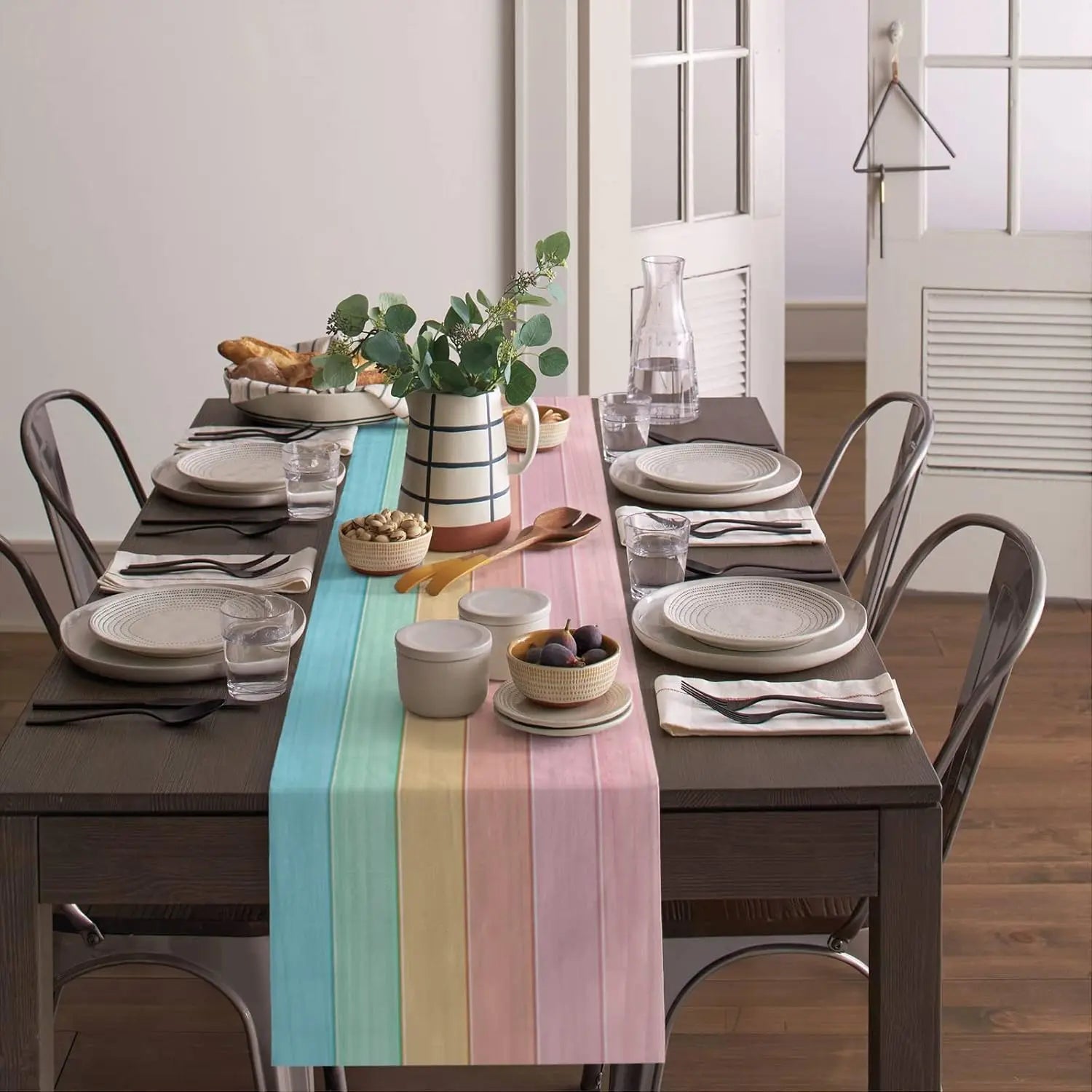 Une scène de table élégante avec une nappe rayée multicolore, de la vaisselle grise, des plantes vertes et un arrangement floral dans un pichet blanc et noir.