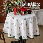 Une nappe de table ronde blanche ornée de motifs d'arbres de Noël et de pompons vert émeraude, avec au centre un bouquet de fleurs rouges, une bougie rouge et une décoration cadeau, créant une ambiance festive de Noël.