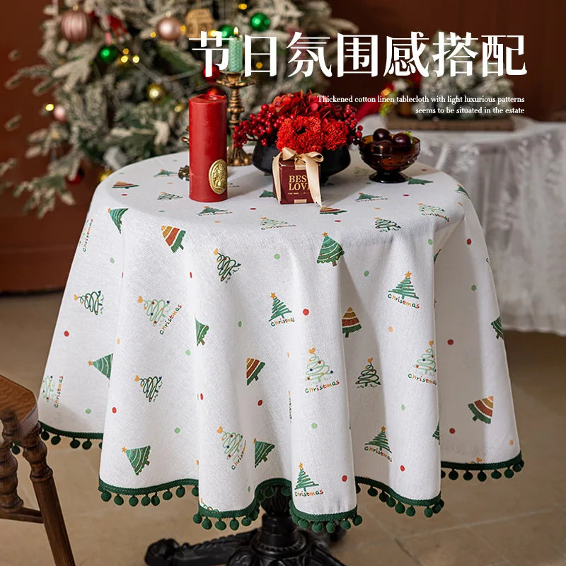 Une nappe de table ronde blanche ornée de motifs d'arbres de Noël et de pompons vert émeraude, avec au centre un bouquet de fleurs rouges, une bougie rouge et une décoration cadeau, créant une ambiance festive de Noël.